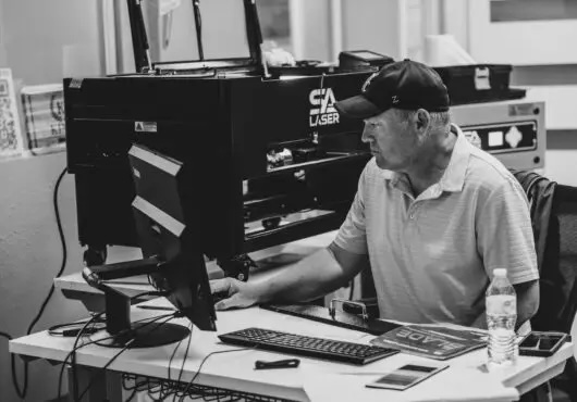 A man sits at a desk while using a desktop computer that is set up next to a SA Laser machine.